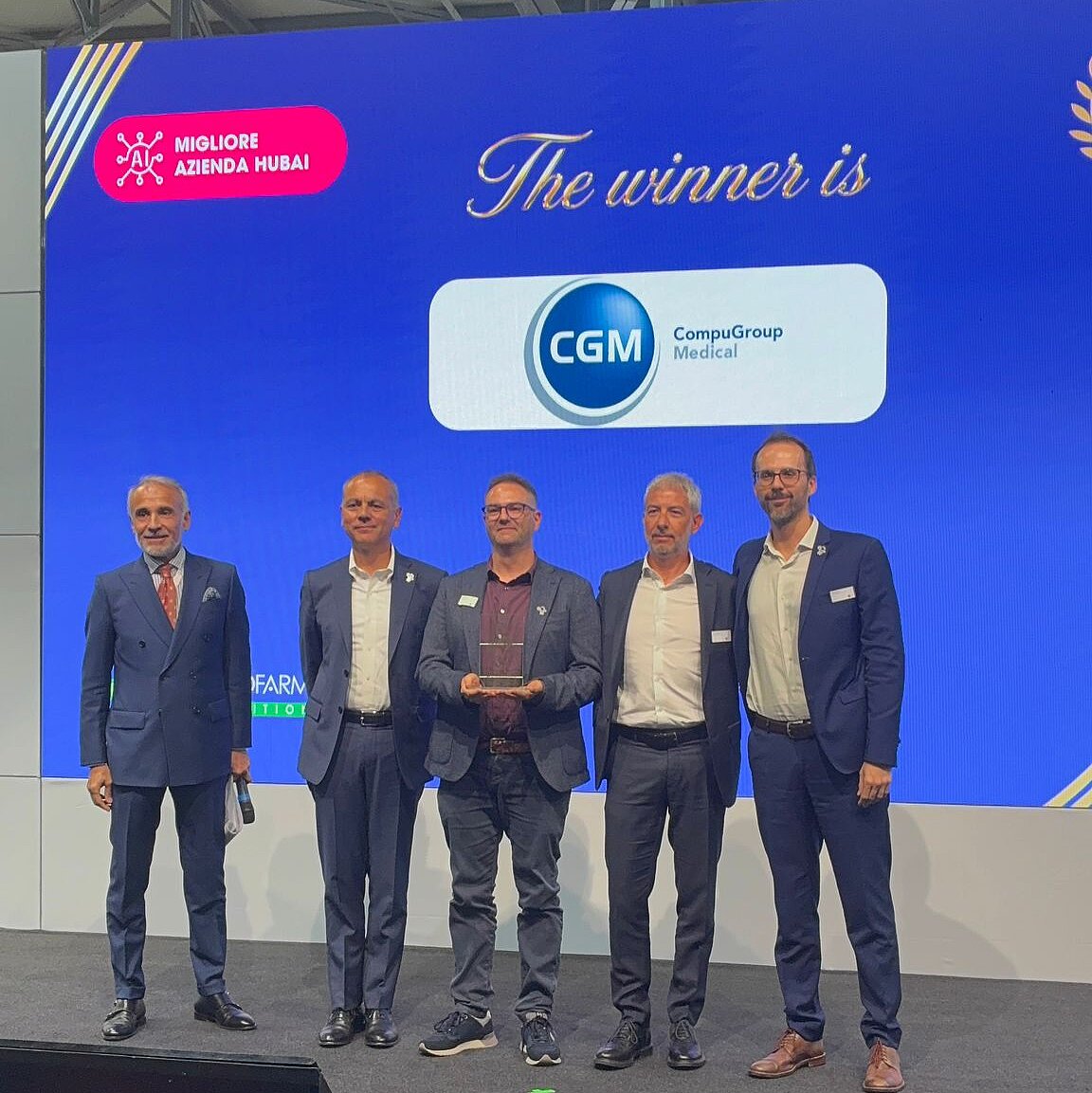 Foto der Preisverleihung in Bologna, CGM-Vertreter von rechts nach links: Dario Rosati (Senior Business Development Manager PCU Pharmacies), Giuseppe Moretti (Area Vice President Product House PCS Europe), Marco Groppo (Product Owner) and Alessandro Avezz Fünf Männer in Anzügen stehen auf einer Bühne vor einer großen blauen Leinwand, auf der CompuGroup Medical als Gewinner der Auszeichnung „Migliore Azienda Hubai“ verkündet wird. Der Mann in der Mitte hält eine gläserne Trophäe.