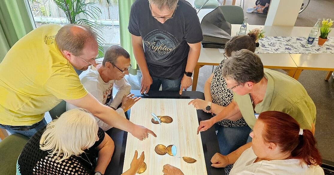 Personen die um einen digitalen Tisch sitzen. Auf dem Tisch ist ein Spiel zu sehen, das mit den fingern bedient wird.