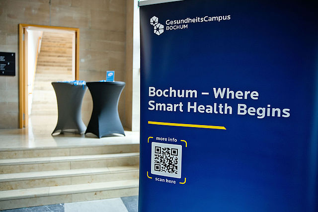 Bochum und Estland vereinbaren engere Zusammenarbeit im Bereich Gesundheitswirtschaft - Donna und der Blitz GmbH Ein blaues Roll-up-Display im Foyer eines Gebäudes. Es zeigt oben das Logo und den Schriftzug „GesundheitsCampus BOCHUM“. Darunter steht der Slogan „Bochum – Where Smart Health Begins“. Im unteren Bereich befindet sich ein QR-Code mit den Hinweisen „more info“ und „scan here“. Im unscharfen Hintergrund sind Stehtische und Treppenstufen zu sehen