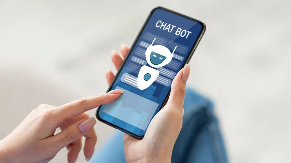 Symbolbild Chatbot © AdobeStock | Prostock Studio Eine Person hält ein Smartphone in den Händen, auf dessen Display eine Chatbot-Benutzeroberfläche mit stilisiertem Roboter-Avatar und dem Schriftzug „CHAT BOT“ zu sehen ist.