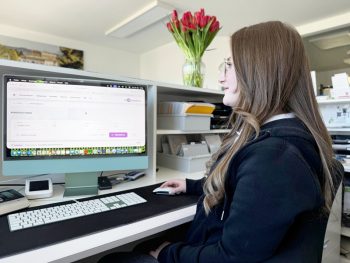 Die Kombination aus KI-gestützter Kommunikation und digitalen Prozessen verbessert Barrierefreiheit und Zusammenarbeit im Praxisalltag. Eine junge Frau mit langen braunen Haaren und Brille sitzt an einem Schreibtisch und arbeitet an einem iMac. Sie schaut auf den Bildschirm, auf dem eine Software-Benutzeroberfläche zu sehen ist, und bedient eine Maus. Im Hintergrund steht eine Vase mit roten Tulpen auf einem Regal.