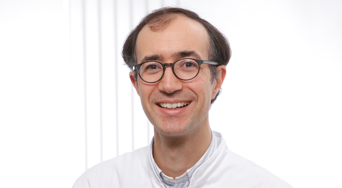 Leitete die internationale Studie, die einen Meilenstein zur weltweit besseren Blutkrebsdiagnostik setzt: Priv.-Doz. Dr. Amin Turki, Leitender Oberarzt der Medizinischen Klinik III – Hämatologie / Onkologie des Marien Hospital Herne. Porträt einer lächelnden Person mit Brille und kurzem, dunklem Haar. Die Person trägt einen weißen Kittel über einem hellen Hemd, was auf einen medizinischen oder wissenschaftlichen Kontext hindeutet. Der Hintergrund ist hell und schlicht gehalten.