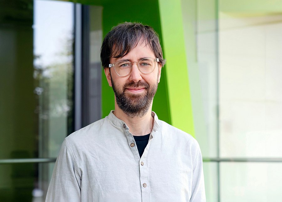 Prof. Dr. Mario Wenzel // Foto Friederike von Heyden Porträt eines lächelnden Mannes mit dunklen Haaren, Vollbart und Brille. Er trägt ein hellgraues Hemd und steht vor einem modernen Gebäude mit Glasfront und grünen Farbelementen.