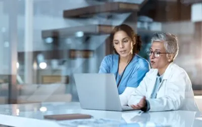 RZV Ausfall-KIS Eine Ärztin und eine Krankenschwester betrachten gemeinsam einen Laptop und besprechen Patienteninformationen in einer modernen medizinischen Einrichtung.