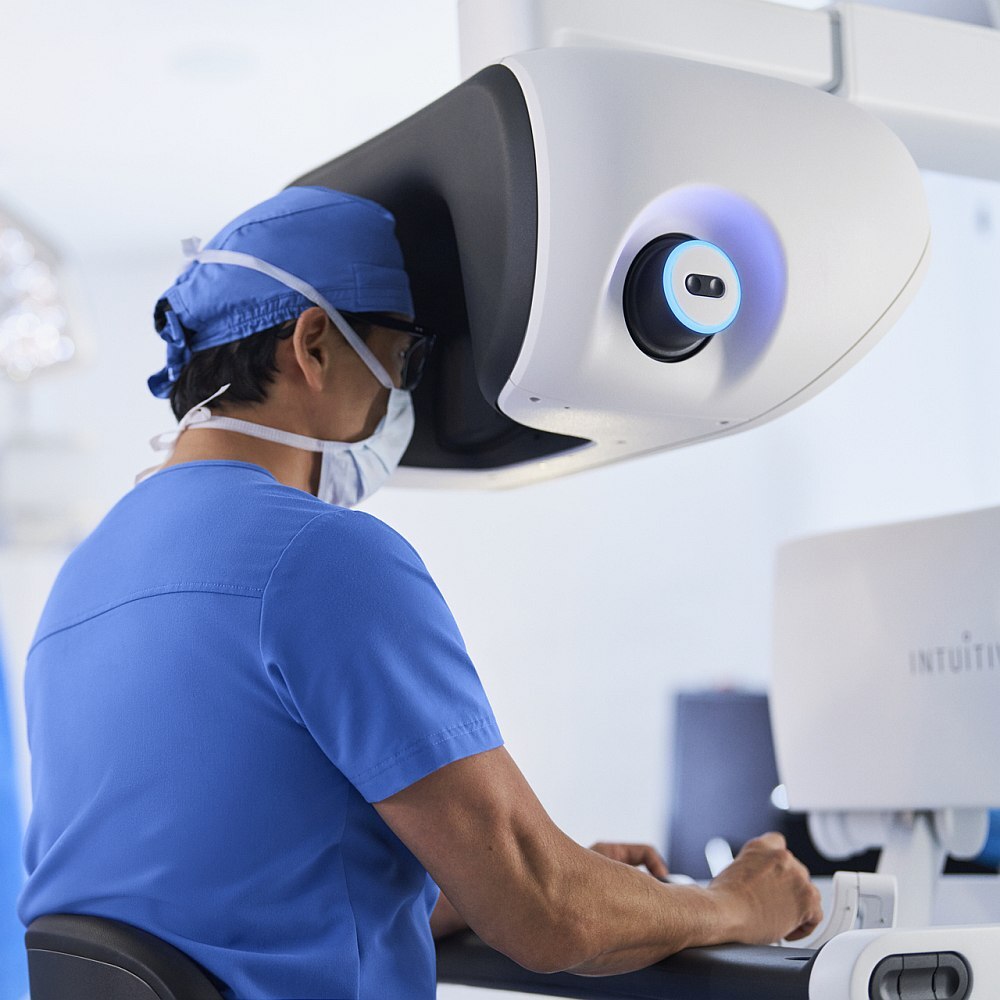 Medizinische Fachkraft in OP-Kleidung bedient ein robotisches Operationssystem. Die Person sitzt an einer Konsole mit Sichtgerät und steuert präzise Instrumente für einen chirurgischen Eingriff in einem modernen Operationssaal.