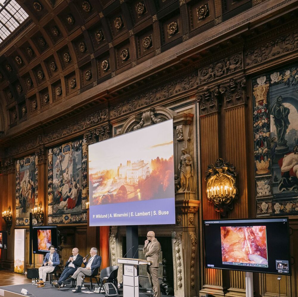 Krupp Robotic Symposium Podiumsdiskussion beim „Krupp Robotic Symposium“ in einem historischen Saal. Ein Moderator steht am Rednerpult, drei Experten sitzen auf der Bühne. Im Hintergrund zeigt eine Leinwand die Namen der Vortragenden (P. Wiklund, A. Minervini, E. Lambert, S. Buse), daneben ein Monitor mit einem medizinischen Endoskopie-Bild.