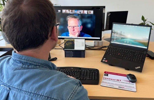 Gemeinsamer Austausch vom NVNR und connect MT Ein Mann mit Brille und Jeanshemd sitzt an einem Schreibtisch und schaut auf einen großen Computermonitor. Der Monitor zeigt eine Videokonferenz mit einem anderen Mann, der in die Kamera blickt. Rechts steht ein aufgeklappter Laptop auf einem Ständer. Auf dem Schreibtisch liegen eine Tastatur, eine Maus, ein Mauspad mit dem Logo „connect MT“ und ein kleiner Tischkalender. Der Hintergrund ist ein helles Büro mit Pflanzen.