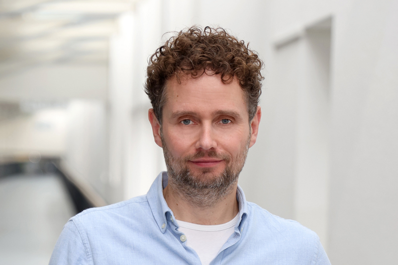 Die Auflösung der Grenzen zwischen physischer Welt und digitaler Informationsverarbeitung sind das Lehrgebiet von Prof. Dr. Peter Detzner, der zum Wintersemester an die Westfälische Hochschule berufen wurde.Foto: WH/Britta Stock Porträt eines Mannes mit lockigem, braunem Haar und einem kurzen Bart. Er blickt direkt in die Kamera und trägt ein hellblaues Hemd über einem weißen T-Shirt. Der Hintergrund zeigt einen hellen, leicht unscharfen Flur.