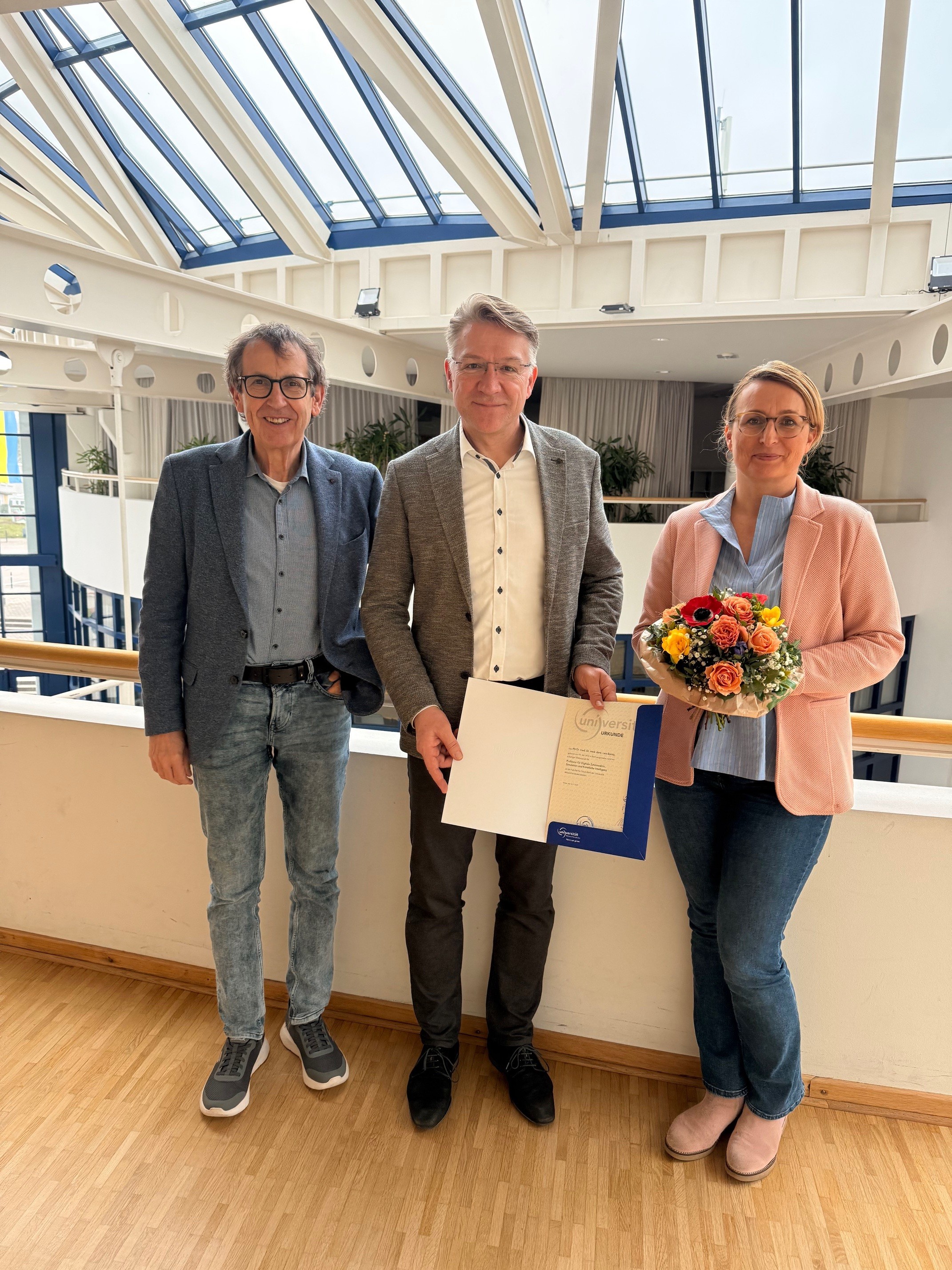 Prof. Dr. Dr. Lars Bonitz wurde auf die Professur für Digitale Zahnmedizin, Simulation und Künstliche Intelligenz berufen. (Foto: UW/H) Drei Personen stehen nebeneinander in einem hellen Innenraum mit Glasdach. Der Mann in der Mitte hält eine Urkunde in einer blauen Mappe, die Frau rechts trägt einen bunten Blumenstrauß. Alle drei blicken freundlich in die Kamera. Das Ambiente wirkt wie ein modernes Büro- oder Universitätsgebäude.